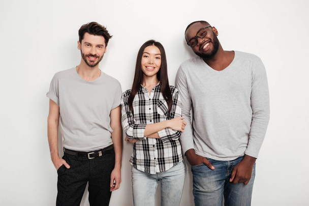 Portrait of three multinational friends standing together and joyfully looking in camera on white background in studio - Photo, Image