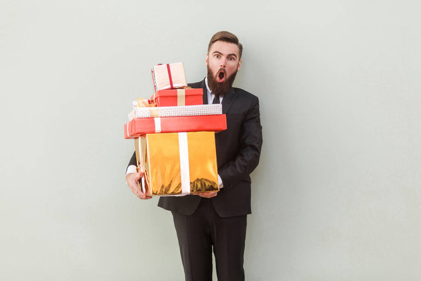 funny man looking at camera with shocked face and holding gift boxes on gray background - Foto, Bild