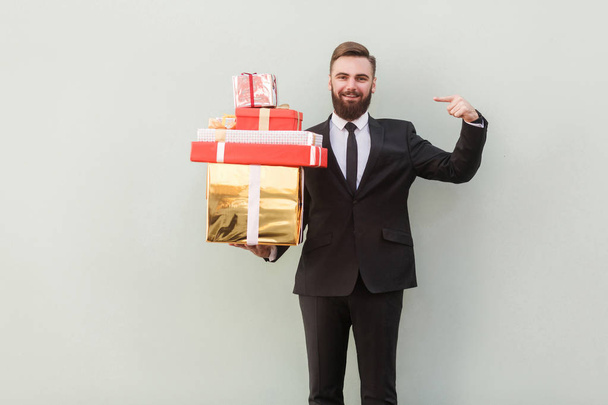 handsome businessman pointing finger at gift boxes and looking at camera on gray background - Фото, изображение