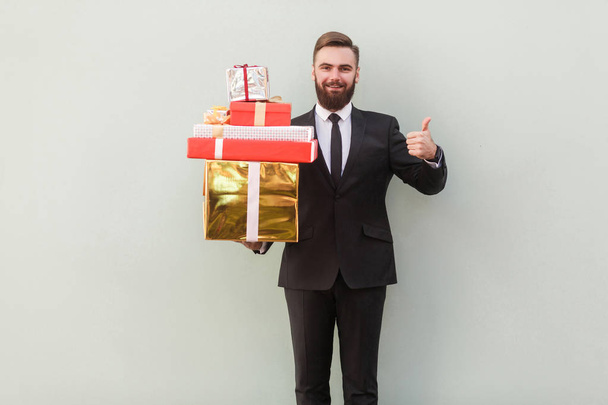 Gelukkig zakenman holding geschenkdozen en duim opdagen op grijze achtergrond - Foto, afbeelding