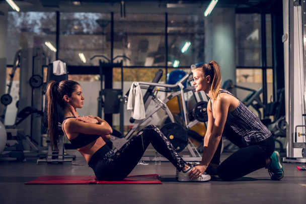 mujer joven haciendo ejercicio mientras su entrenador personal femenino ayuda en el gimnasio
 - Foto, Imagen
