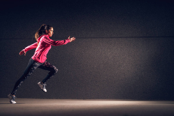 Porträt einer jungen schlanken Frau in Sportkleidung, die gegen eine dunkle Wand läuft - Foto, Bild