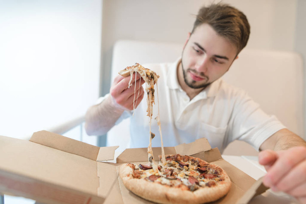A handsome man holds a slender of appetizing pizza in his hands and stares at her with a hungry look. The man eats a hot pizza from a cardboard box. - Photo, Image