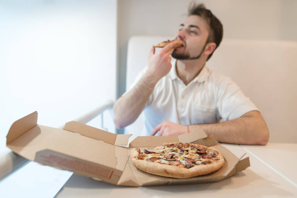 A man in light clothing eats a delicious pizza from a cardboard box. Focus on the cardboard box with pizza. - Photo, image