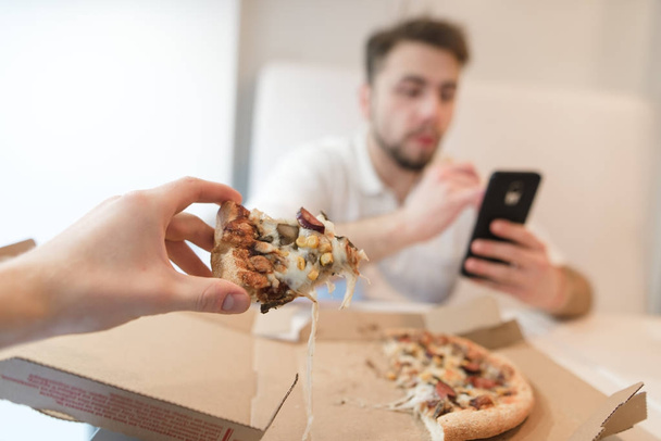 彼の手でピザの作品。若い人たちは、ボックスからピザを食べる。夫は携帯電話を使用し、ピザを食べる. - 写真・画像