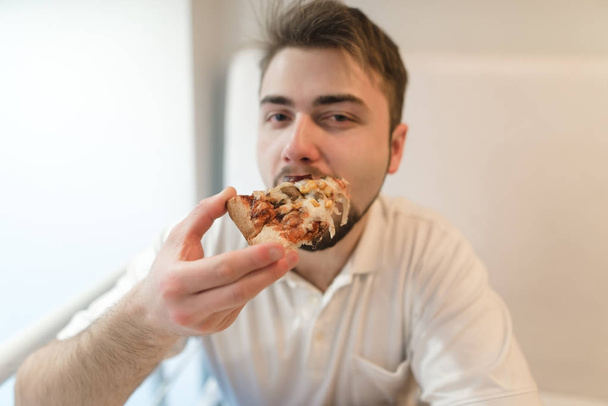 Красивый молодой человек ест аппетитную пиццу и смотрит в камеру. Мальчик с бородой готовит пиццу
. - Фото, изображение