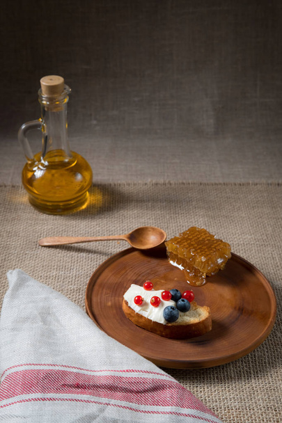 fried bread, toast with yoghurt and berries, bilberries, blueberries and red currants on a wooden plate with honey combs on the background of linen cloth and bottle of oil on the background - Photo, Image