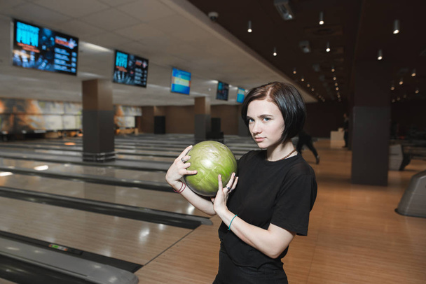 Beautiful young girl playing in a bowling alley with a green ball in her hands. Rest after playing bowling. - Photo, Image