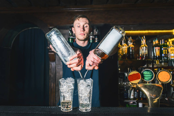 Vista frontal del barman vertiendo alcohol de dos botellas en un cóctel en el bar
 - Foto, Imagen