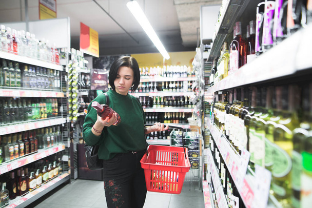 Πορτρέτο του ένα κορίτσι που επιλέγει το αλκοόλ σε ένα σούπερ μάρκετ. Ο σύζυγός της εξετάζει το μπουκάλι κρασί κατά τη στιγμή της ψώνια. Ψώνια στο σούπερ μάρκετ - Φωτογραφία, εικόνα