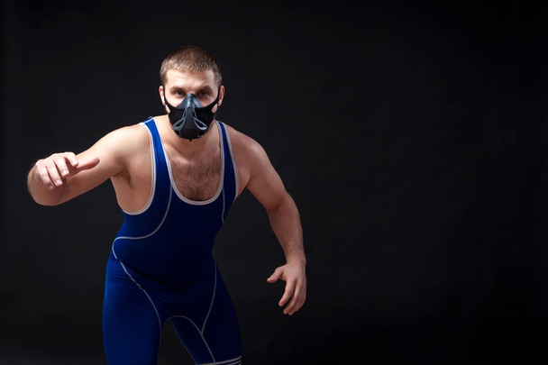 A young dark-haired man fighting Greco-roman wrestling and grappling in a blue   tights  , training mask stands before the start of the  wrestling against a black isolated  background - Photo, Image