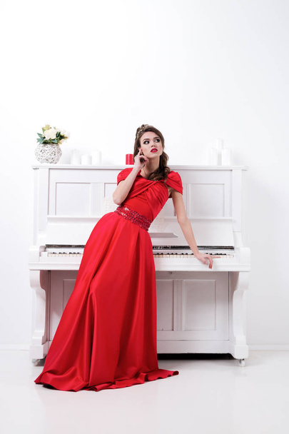 Elegant woman in long red dress standing in white room near pian - Foto, afbeelding