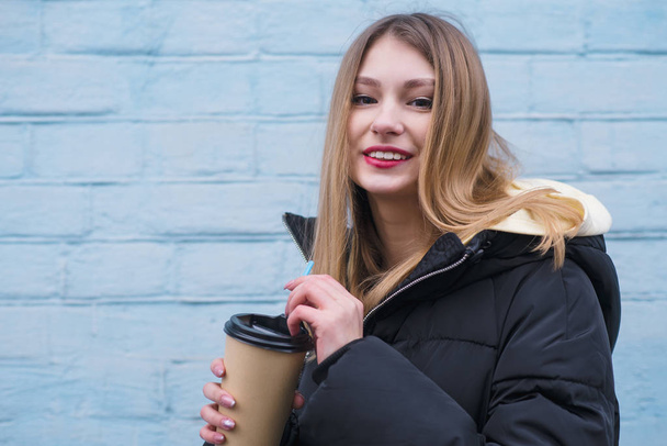 Portret van een lachende jong blond meisje met een kop warme koffie in haar handen tegen de achtergrond van een blauwe muur. - Foto, afbeelding