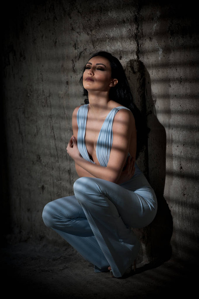 Portrait of beautiful sexy young woman with elegant overall against old wall, urban shot. Attractive young brunette with long hair in white outfit posing fashion in darkness with light stripes.  - Foto, afbeelding