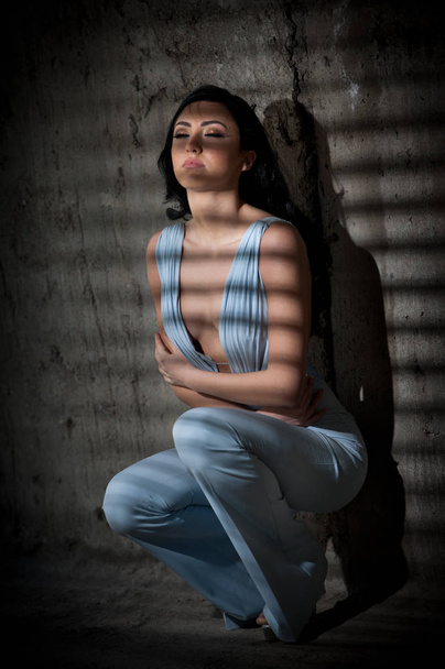 Portrait of beautiful sexy young woman with elegant overall against old wall, urban shot. Attractive young brunette with long hair in white outfit posing fashion in darkness with light stripes.  - Photo, Image