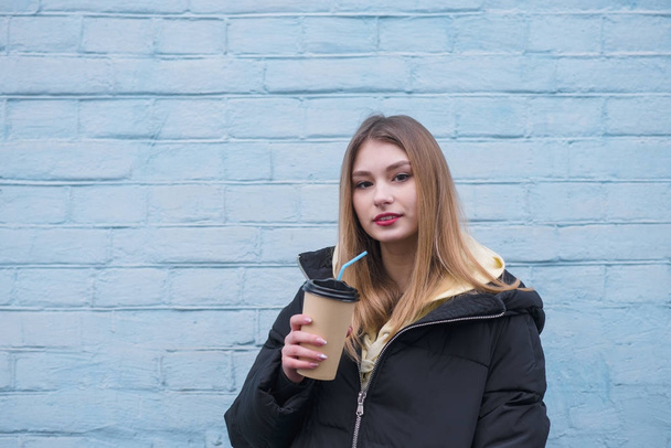 Όμορφη νεαρή ξανθιά κοπέλα πίνοντας τον καφέ στο φόντο της ένα μπλε τοίχο και εξετάζοντας τη φωτογραφική μηχανή. Το κορίτσι σε ένα ζεστό μπουφάν στέκεται στο δρόμο κάτω από τον τοίχο - Φωτογραφία, εικόνα