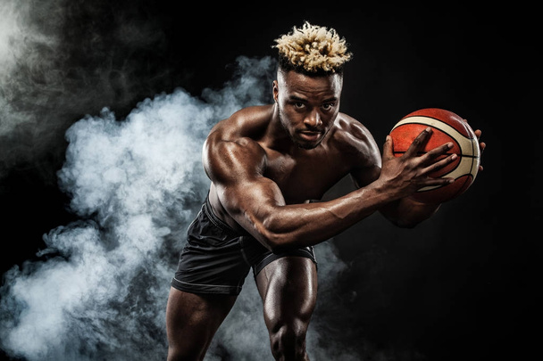 Portrait of afro-american sportsman, basketball player with a ball over black background. Fit young man in sportswear holding ball. - Fotografie, Obrázek