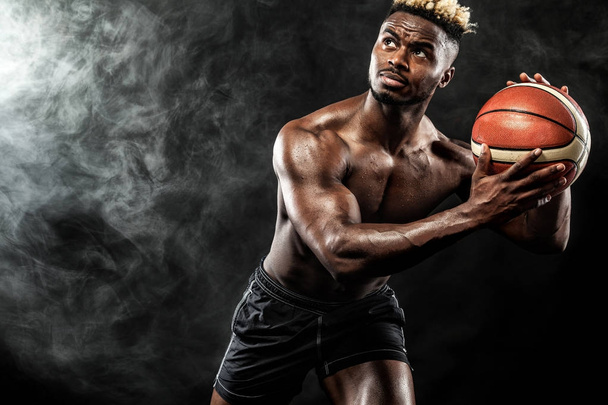 Portrait of afro-american sportsman, basketball player with a ball over black background. Fit young man in sportswear holding ball. - Valokuva, kuva