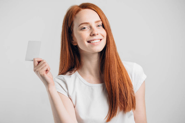 redhead girl holding credit card and smiling on white background. - Photo, Image