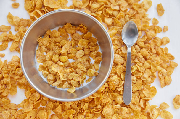 Cornflakes in a metal bowl with milk on a painted white wooden background. Cornflakes scattered on a wooden table with spoon. - Photo, Image