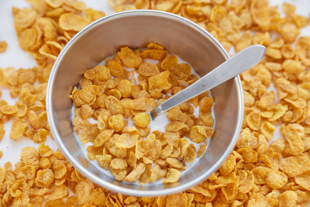 Cornflakes in a metal bowl with milk on a painted white wooden background. Cornflakes scattered on a wooden table with spoon. - Photo, Image