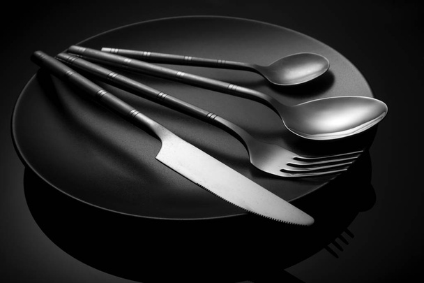 Empty black plate with knife and fork, spoon on a black. - Zdjęcie, obraz