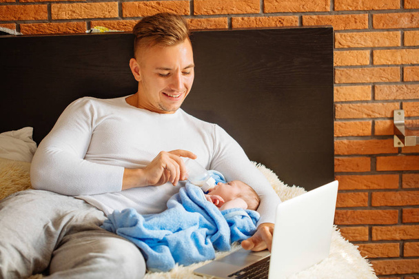 man feeding little baby while lying on bed and working on laptop - Φωτογραφία, εικόνα