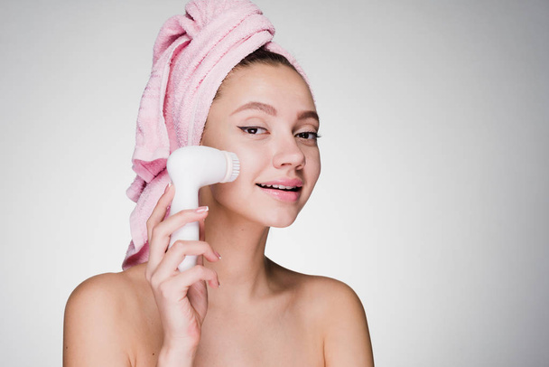 cute smiling girl with a pink towel on her head cleans the skin on her face with an electric brush - Foto, afbeelding
