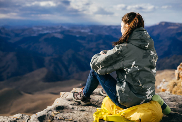 active young girl sits on the edge of a cliff on a yellow backpack, enjoys the mountain nature - Photo, Image