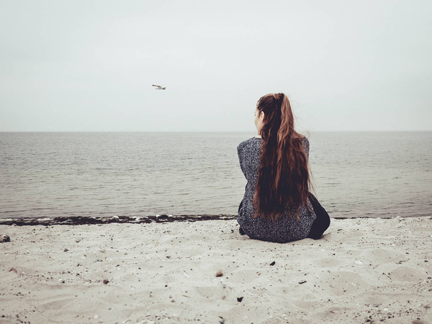 Cute young woman sits on the sand on the beach and looks out into the distance - Fotoğraf, Görsel
