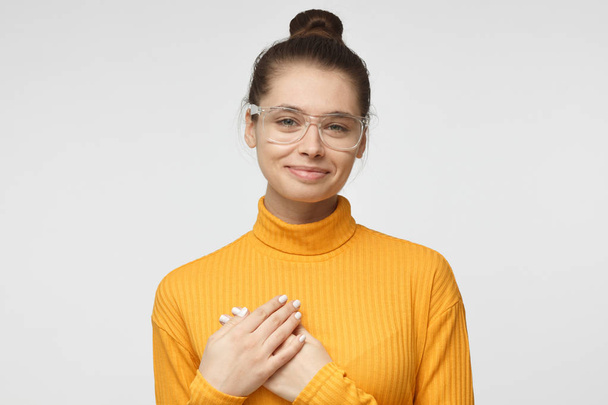 Портрет улыбающейся позитивной молодой женщины держит руки на груди, выражает сочувствие. Добросердечная дружелюбная женщина проявляет доброту, носит горчично-желтый свитер в водолазке
.   - Фото, изображение