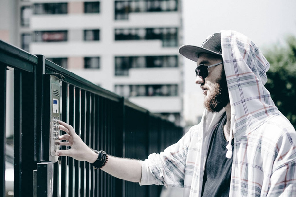 young man with beard in light checkered jacket with hood in a cap and sunglasses picks up the apartment number on the keypad to call the intercom - Foto, Imagem