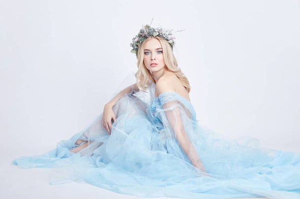 Charming fairy woman in a blue ethereal dress and a wreath on her head on white background, gentle mysterious blonde girl with perfect skin and makeup. Cleanliness, body care and skin. - Foto, immagini