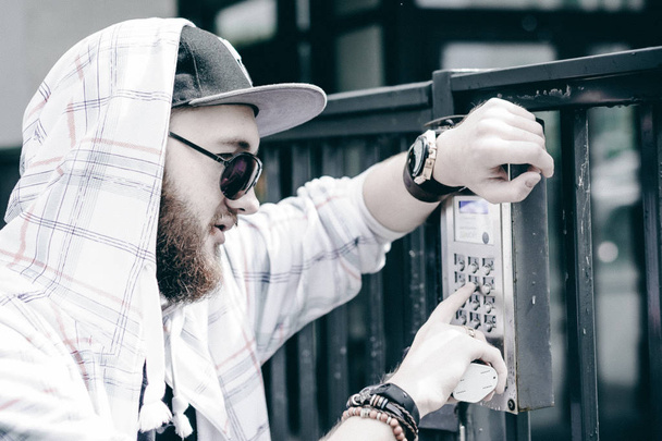 young man with beard in light checkered jacket with hood in a cap and sunglasses picks up the apartment number on the keypad to call the intercom - Foto, immagini