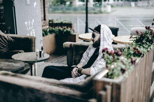young man with beard in light checkered jacket with hood in a cap and sunglasses  sitting on a sofa in a summer cafe - Fotó, kép