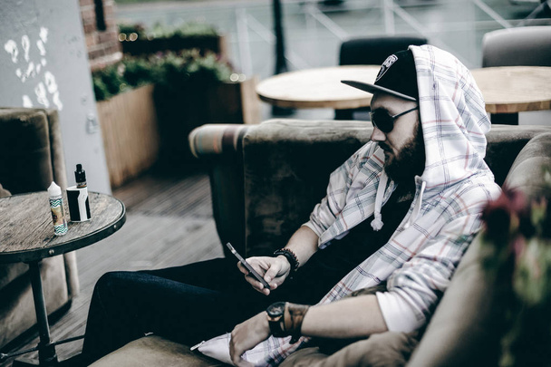 young man with beard in light checkered jacket with hood in a cap and sunglasses  sitting on a sofa in a summer cafe uses a smartphone - Fotoğraf, Görsel