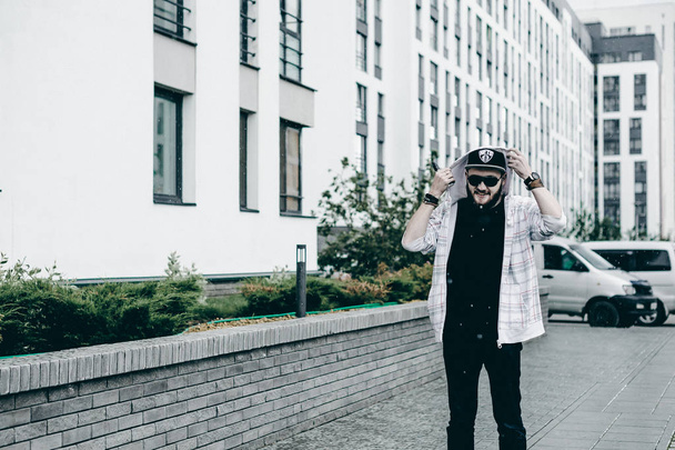 young man with beard in light checkered jacket with hood in a cap and sunglasses walking freely around city smokes an e-cigarette - Fotoğraf, Görsel