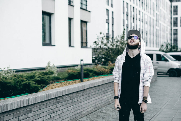 young man with beard in light checkered jacket with hood in a cap and sunglasses walking freely around city smokes an e-cigarette - Foto, Imagem