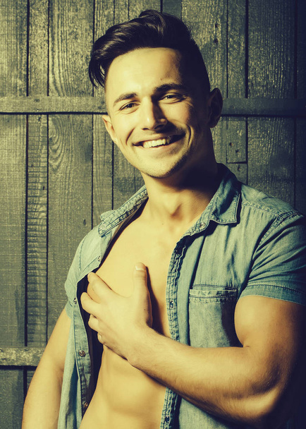 Portrait of happy smiling sexy sensual muscular young macho man with bare torso and stylish hair in jeans shirt indoor on wooden background, vertical picture - Fotó, kép