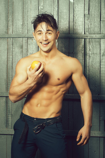 Sexy smiling emotional muscular young man with bare torso holding healthy foor of apple fruit standing indoor on wooden background, vertical picture - Fotó, kép