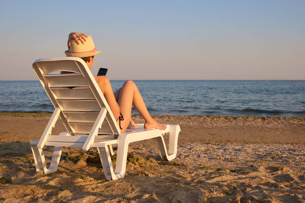 Πίσω προβολή κορίτσι που χρησιμοποιούν smartphone ξαπλωμένη στην ξαπλώστρα παραλίας. - Φωτογραφία, εικόνα