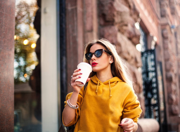 Stylish happy young woman wearing boyfrend jeans, white sneakers bright yellow sweatshirt.She holds coffee to go. portrait of smiling girl in sunglasses she greets friends - Photo, Image