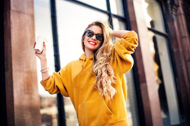 young beautiful blonde drinking coffee walking around the city.mustard sweetshot.,urban backpack , bright red lips Posing against the window of the boutique - Photo, image