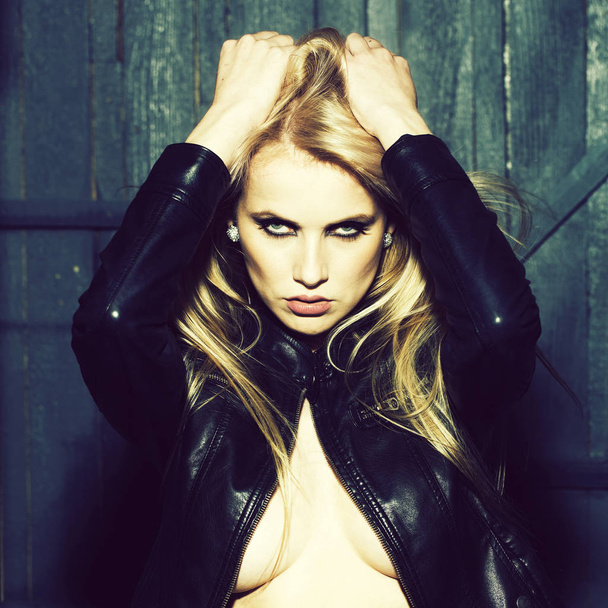 Portrait of one attractive sensual sexy young serious passionate blonde woman touching long hair with hands in leather black jacket in studio on wooden wall background, square picture - Foto, Bild