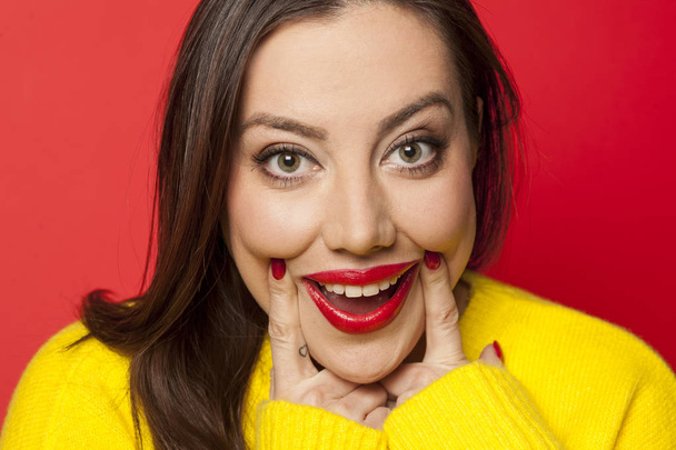 Beautiful woman in yellow sweater forcing her smile with her fingers on a red background - Фото, зображення