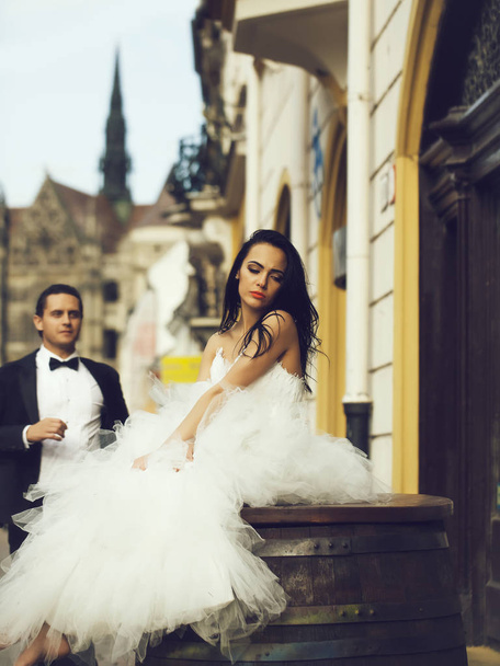 Beautiful bride brunette woman with long hair in fashion white wedding dress sits on wooden barrel and waits for groom on streetscape - Φωτογραφία, εικόνα
