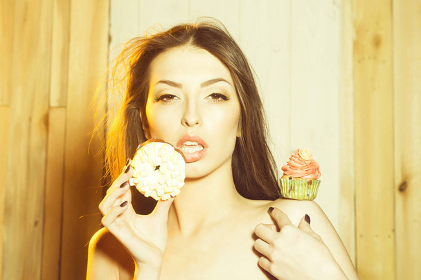 Closeup portrait of one beautiful sensual young woman with bare shoulders near tasty fresh homemade cup cake with orange cream with flower icing in green paper and doughnut  indoor, horizontal photo - Zdjęcie, obraz