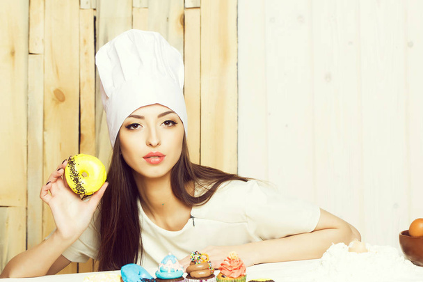 One smiling cute young female cook in professional white uniform of hat and apron cooking sweet cupcake and doughnut desserts of flour and eggs in kitchen on wooden background, horizontal picture - Photo, Image