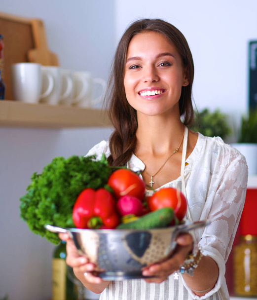 Smiling young woman holding vegetables standing in kitchen - Photo, image