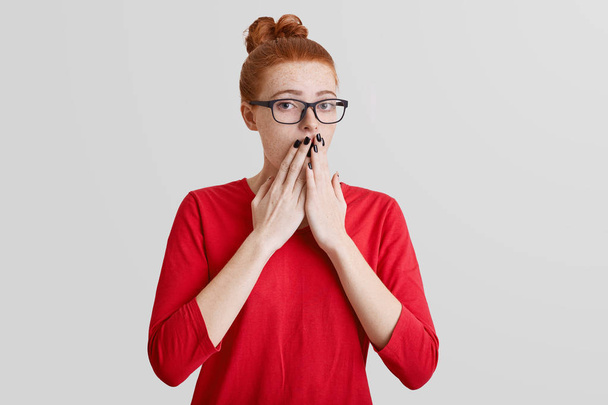 Startled frightened ginger woman covers mouth with hand, being afraid of something, wears spectacles and red sweater, poses alone against white background. Human reaction and feeling concept - Foto, Bild